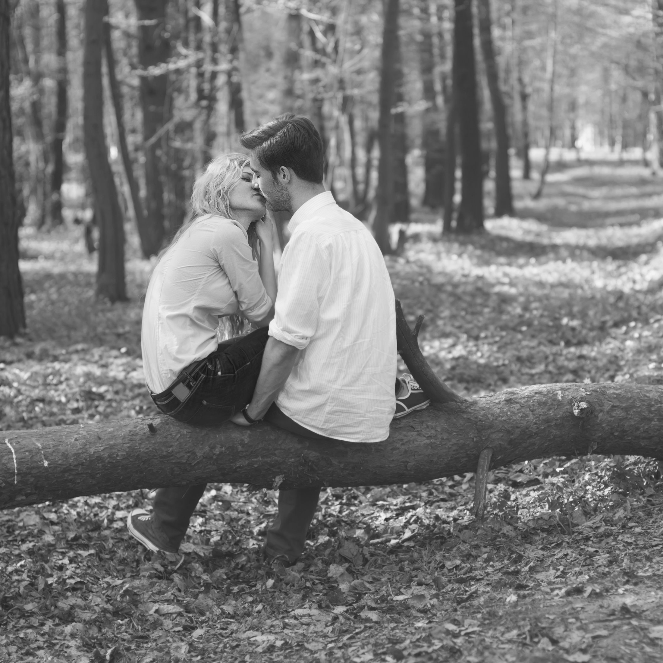 Black and white photo of a couple kissing on a fallen tree in the middle of a forest