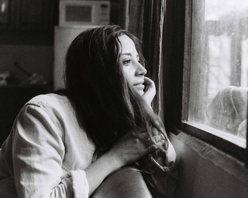 A woman with long, dark hair rests her chin on her hand by the window, quietly focusing on her partner's patterns.